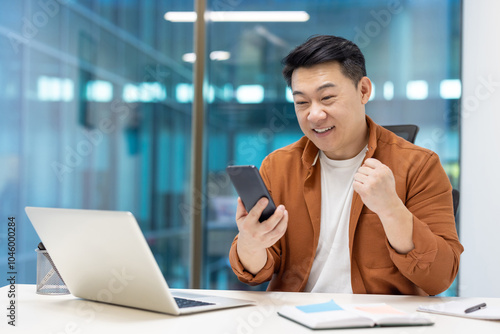 Asian businessman showing excitement looking at phone at office desk. Celebrating success in professional environment with modern tech tools around. Positive energy and motivation.