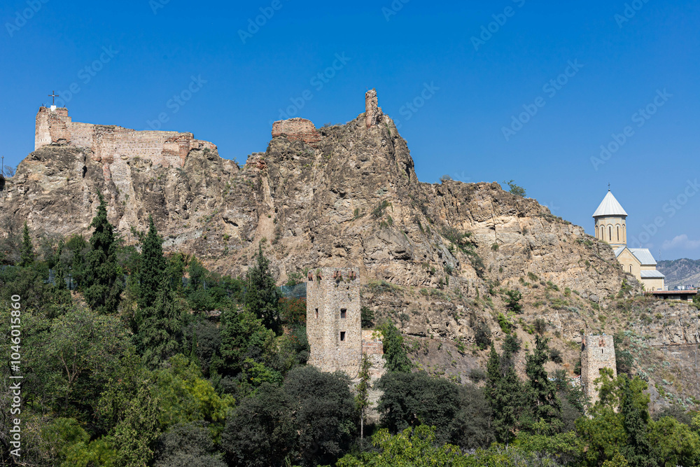view of houses and towers in a mountain or rock in Tbilisi Botanical Garden, Georgia against blue sky, church in the distance view of houses and towers in a mountain or rock in Tbilisi Botanical Garden, Georgia against blue sky, church in the distance