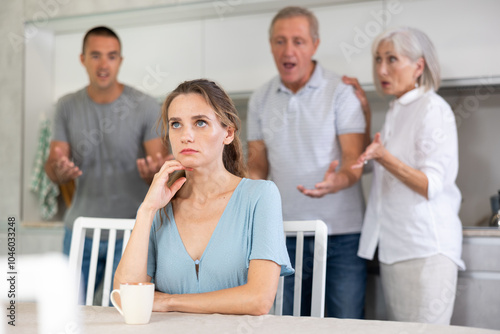 Domestic quarrel - offended young woman sits at the table while parents and young husband yell at her photo