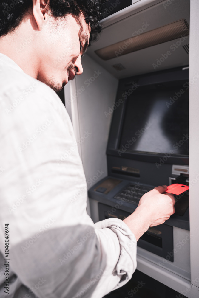 Man inserting a red bank card into an ATM, captured from a close side angle. The screen is dark, emphasizing the action of the user interacting with the cash machine Man inserting a red bank card into an ATM, captured from a close side angle. The screen is dark, emphasizing the action of the user interacting with the cash machine