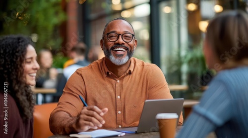 A cheerful man sits at a cafe table, engaged with two friends, holding a pen and notebook, with coffee and a laptop, enjoying a business discussion.