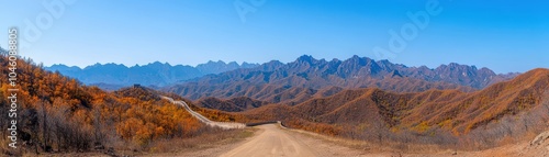 Stunning panoramic view of mountains and dirt road under clear blue sky, showcasing vibrant autumn foliage.