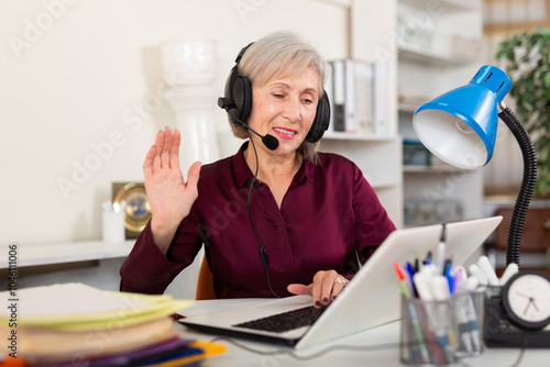 Portrait of smiling mature female employee in earphones looking at laptop screen at workplace in office photo