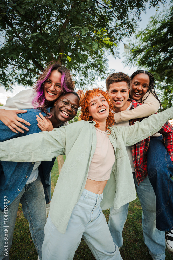 Obraz premium Vertical. Group of joyful young friends having fun outdoors, embracing and laughing together in a park. Energetic, carefree and multiracial people enjoying a lively and playful moment of friendship
