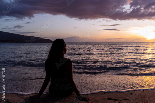 Una mujer sentada en la playa al atardecer, mirando el agua A woman sits on the beach at sunset, looking out at the water 