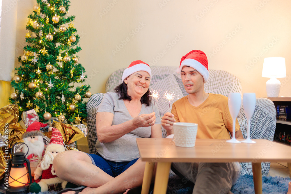Fototapeta premium Brazilian mother and adult son celebrating Christmas at home, sitting by a decorated tree, holding sparklers and wearing Santa hats in a warm, joyful moment