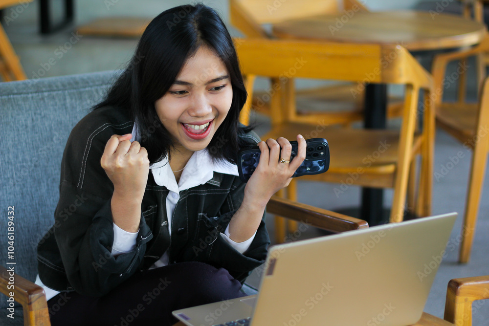 cheerful asian woman holding smartphone sitting in cafe raising fist showing winning gesture while using laptop working remotely cheerful asian woman holding smartphone sitting in cafe raising fist showing winning gesture while using laptop working remotely