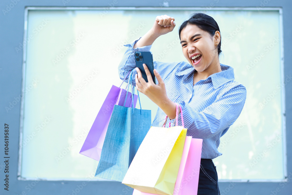 happy asian woman carrying shopping bag and holding smartphone with raised fist celebrating victory gesture walking in outdoors with empty billboard background happy asian woman carrying shopping bag and holding smartphone with raised fist celebrating victory gesture walking in outdoors with empty billboard background