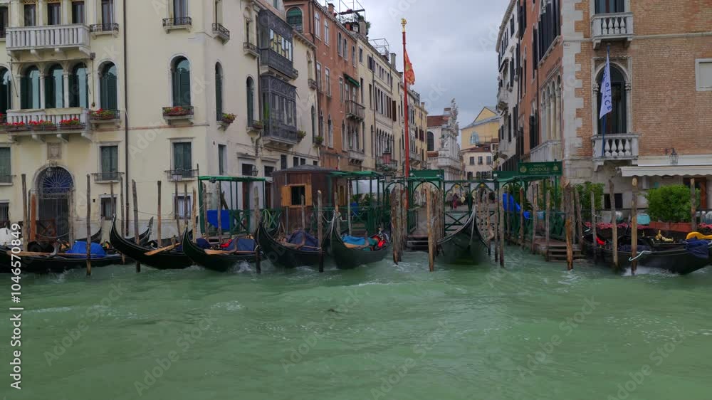 A row of gondolas is moored along the edge of historic buildings in Venice, with calm green waters and a cloudy sky above. A row of gondolas is moored along the edge of historic buildings in Venice, with calm green waters and a cloudy sky above.