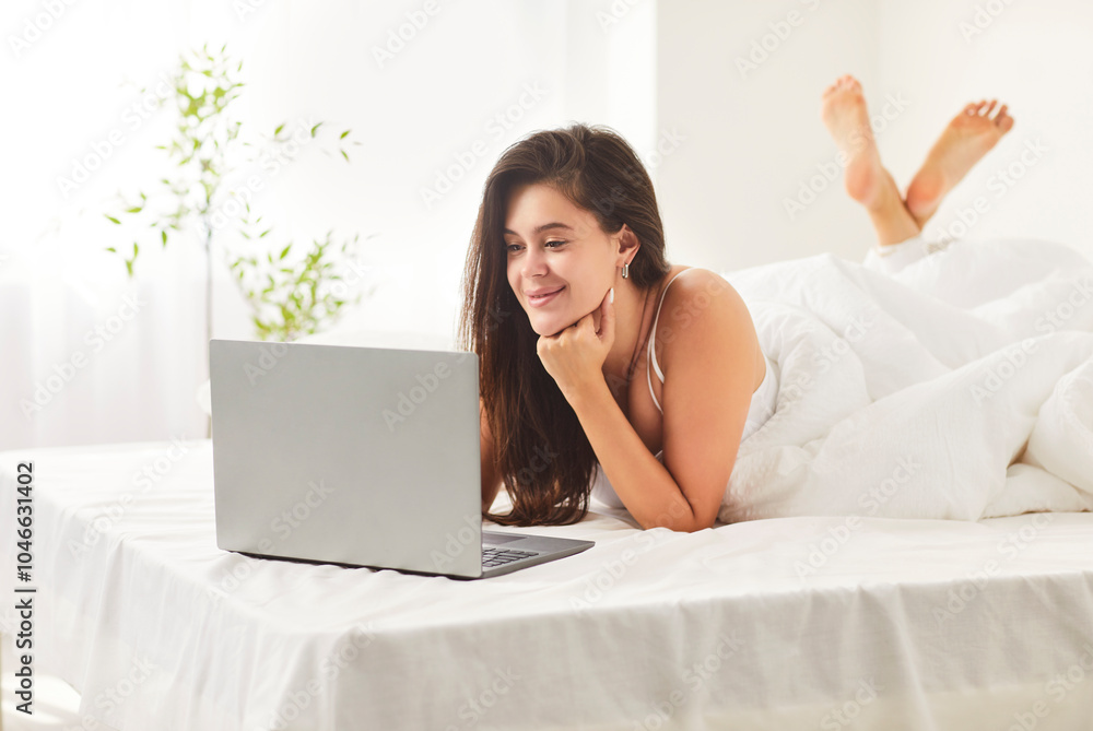 Pretty young woman lying in comfortable bed under white duvet with laptop. Happy smiling caucasian girl surfing internet, watching movies, enjoying leisure time in sunlit bedroom during weekend. Pretty young woman lying in comfortable bed under white duvet with laptop. Happy smiling caucasian girl surfing internet, watching movies, enjoying leisure time in sunlit bedroom during weekend.