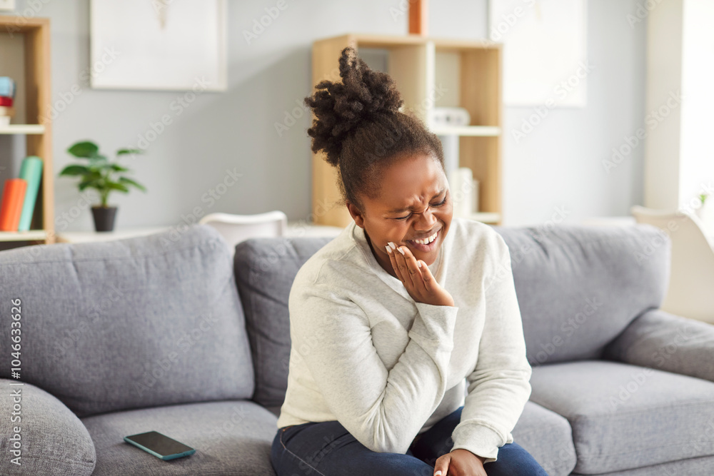Portrait of a young upset african american woman sitting on sofa in living room at home suffering from toothache. Sad girl holding her cheek in dental pain. Health care and dentistry concept. Portrait of a young upset african american woman sitting on sofa in living room at home suffering from toothache. Sad girl holding her cheek in dental pain. Health care and dentistry concept.