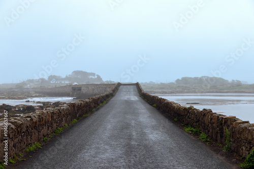 A narrow stone bridge in Connemara, Ireland, extending into the misty distance under an overcast sky, with wet road surface reflecting the foggy ambiance of a quiet, rural landscape photo