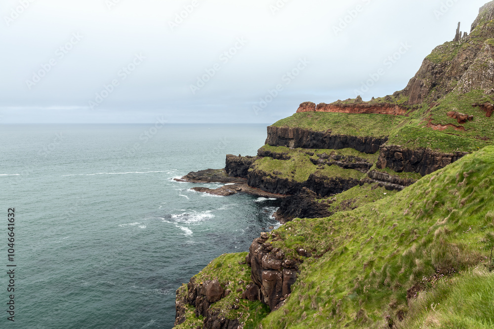 Coastal cliffs at Giant Causeway on a cloudy day. The ocean merges with the sky, while the textured cliffs and bright green grass contrast against the soft light Coastal cliffs at Giant Causeway on a cloudy day. The ocean merges with the sky, while the textured cliffs and bright green grass contrast against the soft light