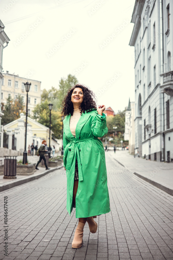 Fototapeta premium Beautiful stylish young woman with curly dark hair posing in a green trench coat in an urban context. Street fashion and style.