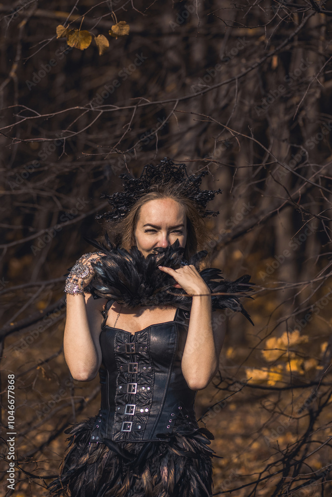 Naklejka premium A young woman in the form of a witch and sorceress, standing in a dark forest, wearing a black dress with feathers. A beautiful gothic queen works wonders in the autumn forest on Halloween.