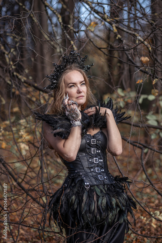 Naklejka premium A young woman in the form of a witch and sorceress, standing in a dark forest, wearing a black dress with feathers. A beautiful gothic queen works wonders in the autumn forest on Halloween.