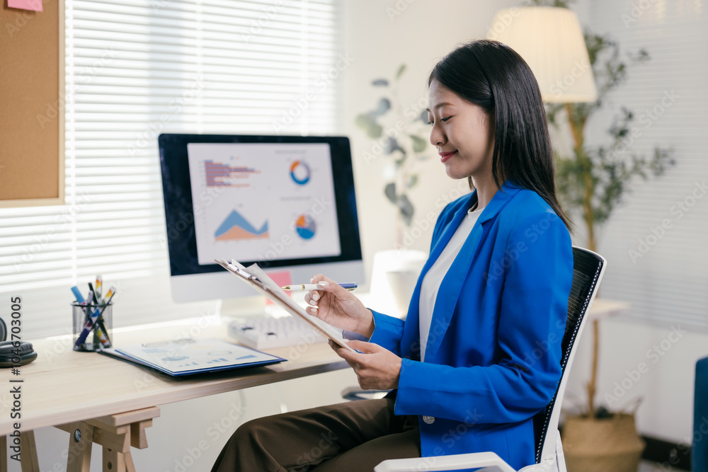 Confident young asian businesswoman in a modern office, analyzing financial data on her computer, taking notes and smiling Confident young asian businesswoman in a modern office, analyzing financial data on her computer, taking notes and smiling