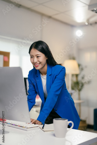 Young asian businesswoman smiles while working on her computer in a bright office with a blue interior and natural light from a window photo