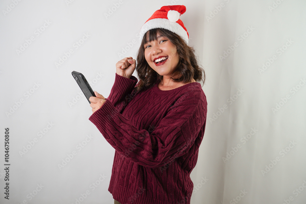 Naklejka premium Excited young asian woman wearing santa clause hat and red long sleeved sweater is using smartphone, isolated over white background. Concept for Christmas Holiday and New Year Party