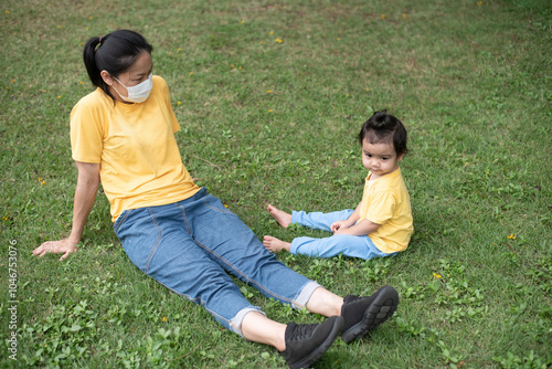 Asian mother wear face mask and daughter sitting together, relaxing, resting on  greensward