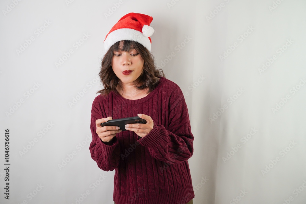 Excited young asian woman wearing santa clause hat and red long sleeved sweater is using smartphone, isolated over white background. Concept for Christmas Holiday and New Year Party Excited young asian woman wearing santa clause hat and red long sleeved sweater is using smartphone, isolated over white background. Concept for Christmas Holiday and New Year Party