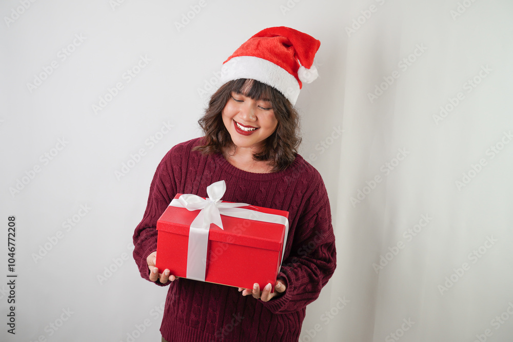 Excited young asian woman wearing santa clause hat and red long sleeved sweater is carrying large gift box with ribbon, isolated over white background. Concept for Christmas Holiday and New Year Party Excited young asian woman wearing santa clause hat and red long sleeved sweater is carrying large gift box with ribbon, isolated over white background. Concept for Christmas Holiday and New Year Party