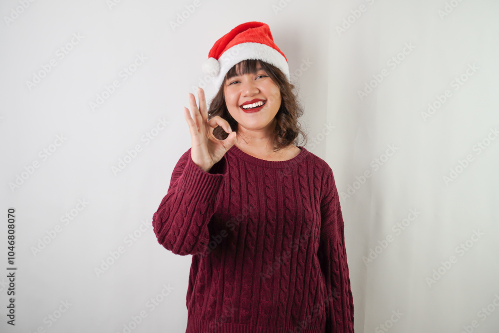 Excited young asian woman wearing santa clause hat and red long sleeved sweater is showing OK Hand Gestures, isolated over white background. Concept for Christmas Holiday and New Year Party Excited young asian woman wearing santa clause hat and red long sleeved sweater is showing OK Hand Gestures, isolated over white background. Concept for Christmas Holiday and New Year Party