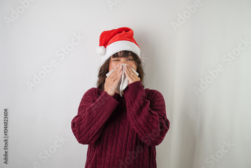 Sick young asian woman wearing santa clause hat and red long sleeved sweater is sneezing and covering using tissue, isolated over white background. Concept for Christmas Holiday and New Year Party