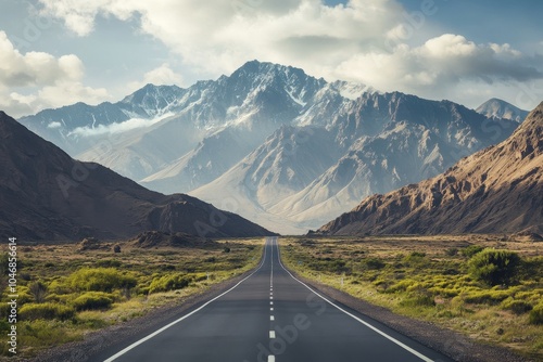 A straight paved road leading to a snow-capped mountain range under a blue sky with clouds.