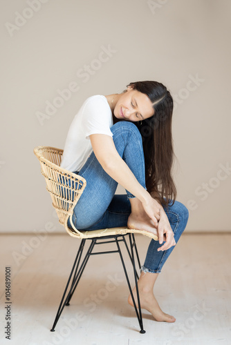 Portrait of confident beautiful woman with long brown hair, wearing casual clothes, sitting on chair in tight jeans and white t-shirt, studio background photo