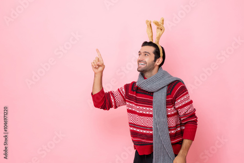 Christmas studio shot portrait of smiling Caucasian man wearing sweater and fancy reindeer headband poiting hand up to copy space in pink color isolated background photo