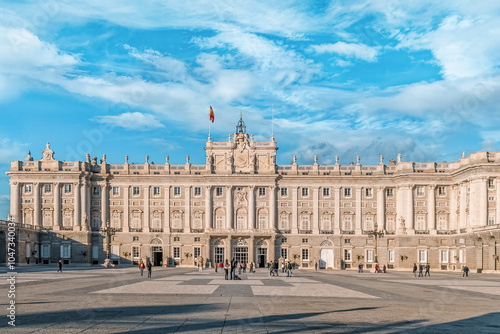 The Royal Palace of Madrid stands majestically under a bright blue sky, its neoclassical facade showcasing detailed columns, stone carvings, and grand windows. Madrid, Spain - November 30, 2021