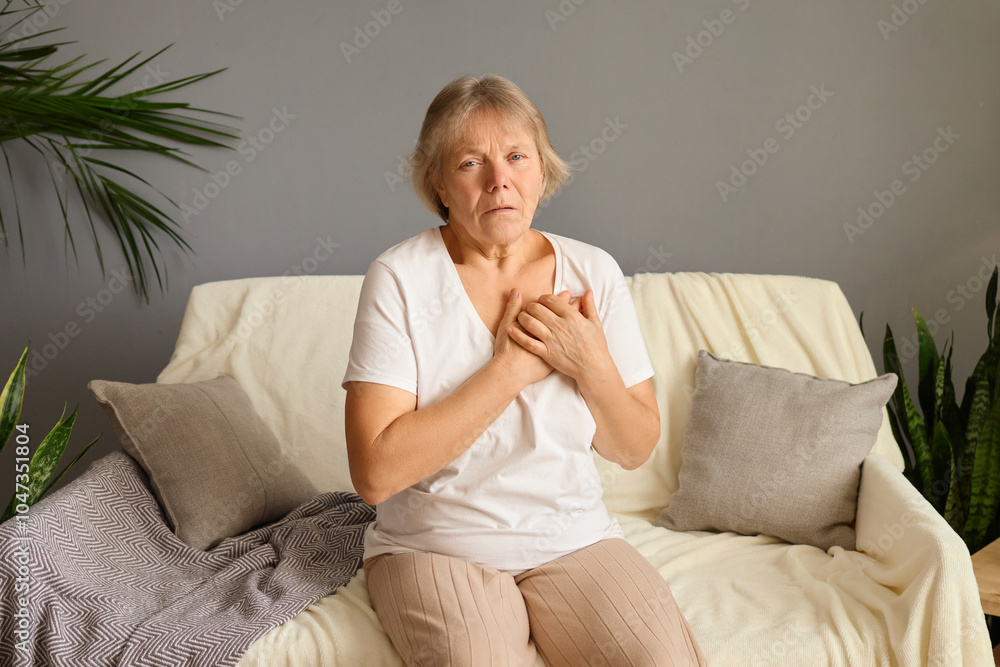 Mature woman with hand over heart expressing severe pain and exhaustion suffering from acute chest discomfort or cardiac issue appearing distressed and uncomfortable sitting alone on couch at home Mature woman with hand over heart expressing severe pain and exhaustion suffering from acute chest discomfort or cardiac issue appearing distressed and uncomfortable sitting alone on couch at home