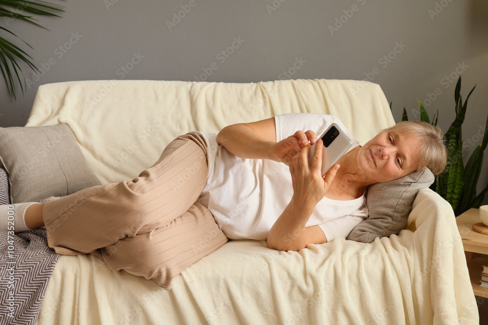 Mature woman lying on sofa at home scrolling through smartphone happily browsing online shopping and checking social media using modern technology and enjoying her retired life Mature woman lying on sofa at home scrolling through smartphone happily browsing online shopping and checking social media using modern technology and enjoying her retired life