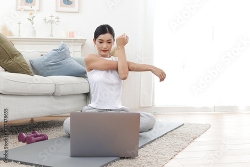 Asian woman stretching body on yoga mat in living room. Happy female working out while watch training tutorial vdo on laptop computer at home. Sporty girl doing exercise. Healthy and sport hobby photo