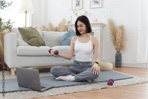 Asian woman lift dumbbell, watch training tutorial vdo on laptop computer. Happy female working out on yoga mat in living room at home. Beautiful sporty girl doing workout. Healthy and sport hobby. photo