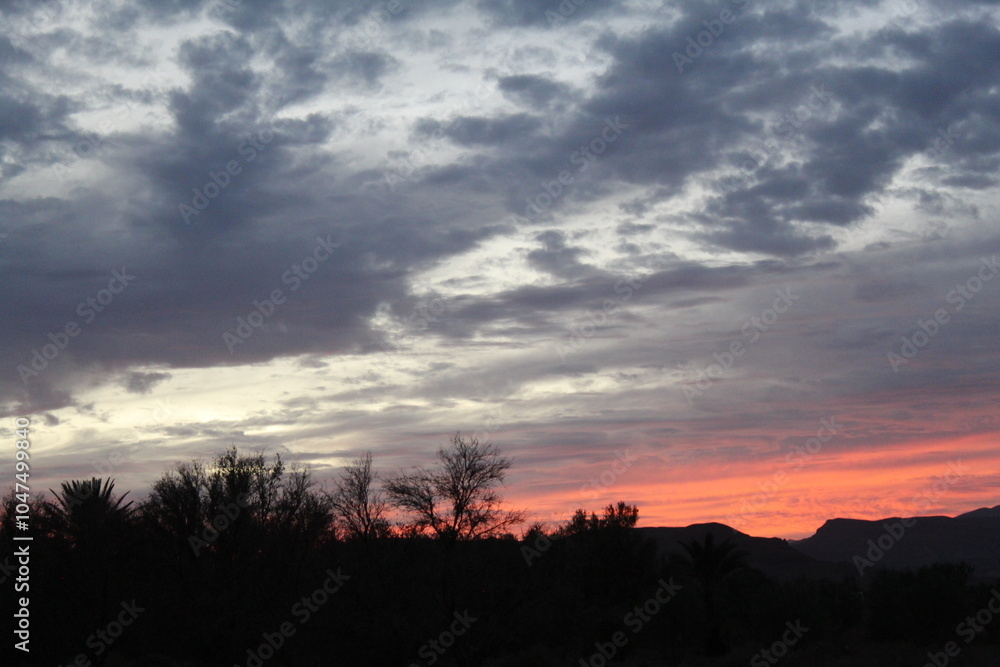 A stunning sunset unfolds like a work of art, with dramatic skies and scattered dark clouds. Vibrant orange and red hues highlight the sun, while tree and mountain silhouettes stand against this maste A stunning sunset unfolds like a work of art, with dramatic skies and scattered dark clouds. Vibrant orange and red hues highlight the sun, while tree and mountain silhouettes stand against this maste