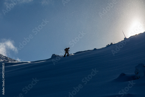 A solitary figure climbs through deep snow, heading toward the mountain's peak as sunlight glows behind.