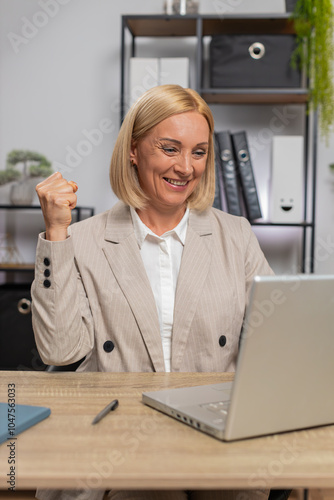 Happy Caucasian woman celebrating success reading great news on laptop while sitting at desk at workplace in home office. Smiling businesswoman satisfied with nice notification, shows yes gesture. photo