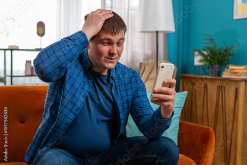 Sad displeased man using smartphone browsing, loses becoming surprised sudden lottery game results, bad news, fortune loss, fail, fraud, virus. Young Caucasian guy on couch in living room at home. photo