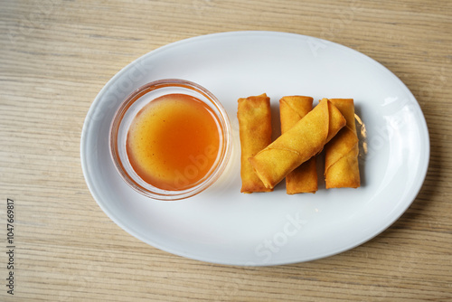 Small crispy fried spring rolls and a glass bowl with sweet and sour sauce on a white plate and a wooden table, appetizer in a Chinese restaurant, high angle view from above, copy spac photo