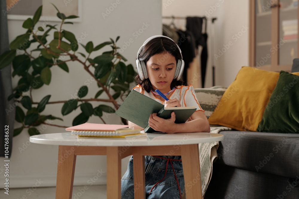 Young girl wearing headphones studying in cozy room with plants and books surrounding her, focused on writing in notebook while seated at table Young girl wearing headphones studying in cozy room with plants and books surrounding her, focused on writing in notebook while seated at table