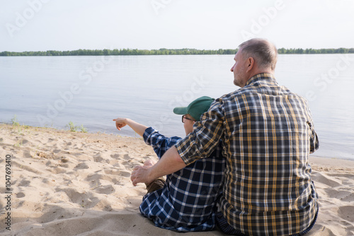 Obraz na płótnie Father's day concept. Rear view of father and son sitting together on the river bank and looking at the water. w salonie