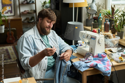 Smiling man sitting by table with sewing machine and stitching detail to denim jacket or jeans while upcycling secondhand apparel photo