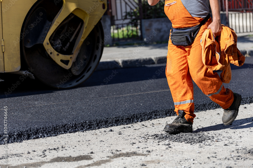 Construction worker in orange uniform walks along freshly laid asphalt on a sunny day in an urban area Construction worker in orange uniform walks along freshly laid asphalt on a sunny day in an urban area