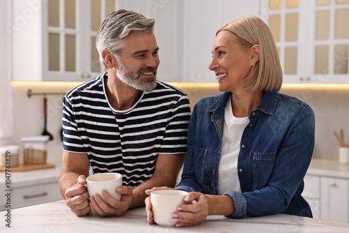 Happy middle aged couple with cups of drink at white marble table in kitchen