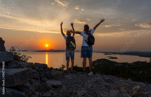 Mother and son standing on rocky mountain trail at sunset, overlooking vast sea and distant islands. Concept of family adventure, outdoor exploration, quality time in nature. High quality photo