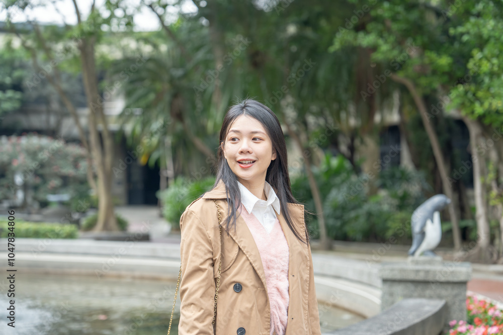 Fototapeta premium A long-haired Taiwanese woman in her 20s wearing a beige trench coat spends her winter afternoon around a fountain in a park in Xinyi District, Taipei, Taiwan, on January 30, 2024.