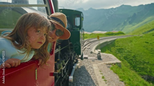 A mother and her little daughter ride in the famous red rotary steam train leaving the town of Brienz in the Swiss Alps along the alpine meadows to the top of a mountain in summer. Switzerland