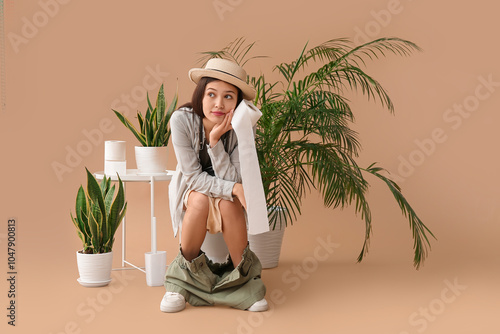 Young Asian traveler with paper roll sitting on toilet bowl against beige bac...